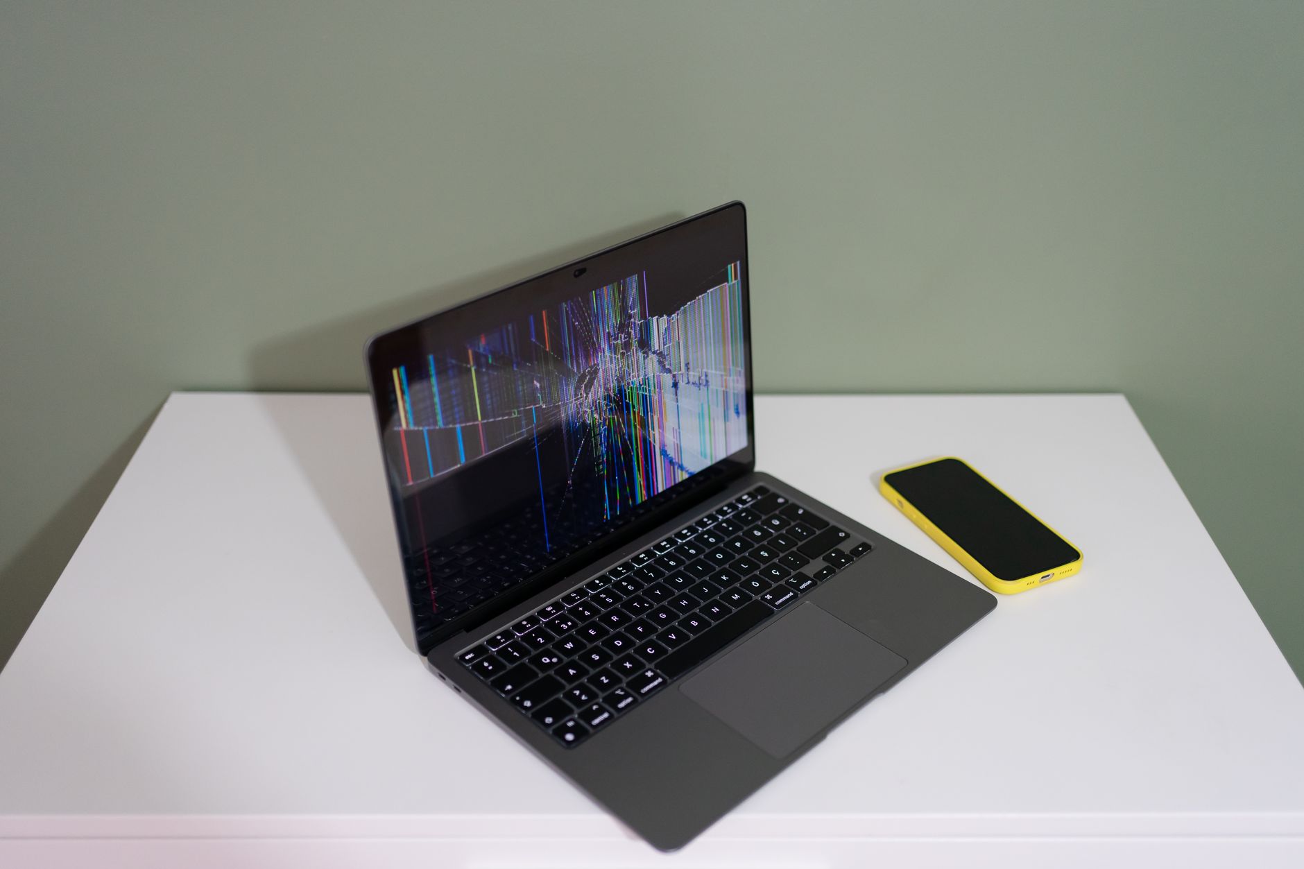 A damaged laptop screen with colorful lines and a yellow smartphone on a white desk.